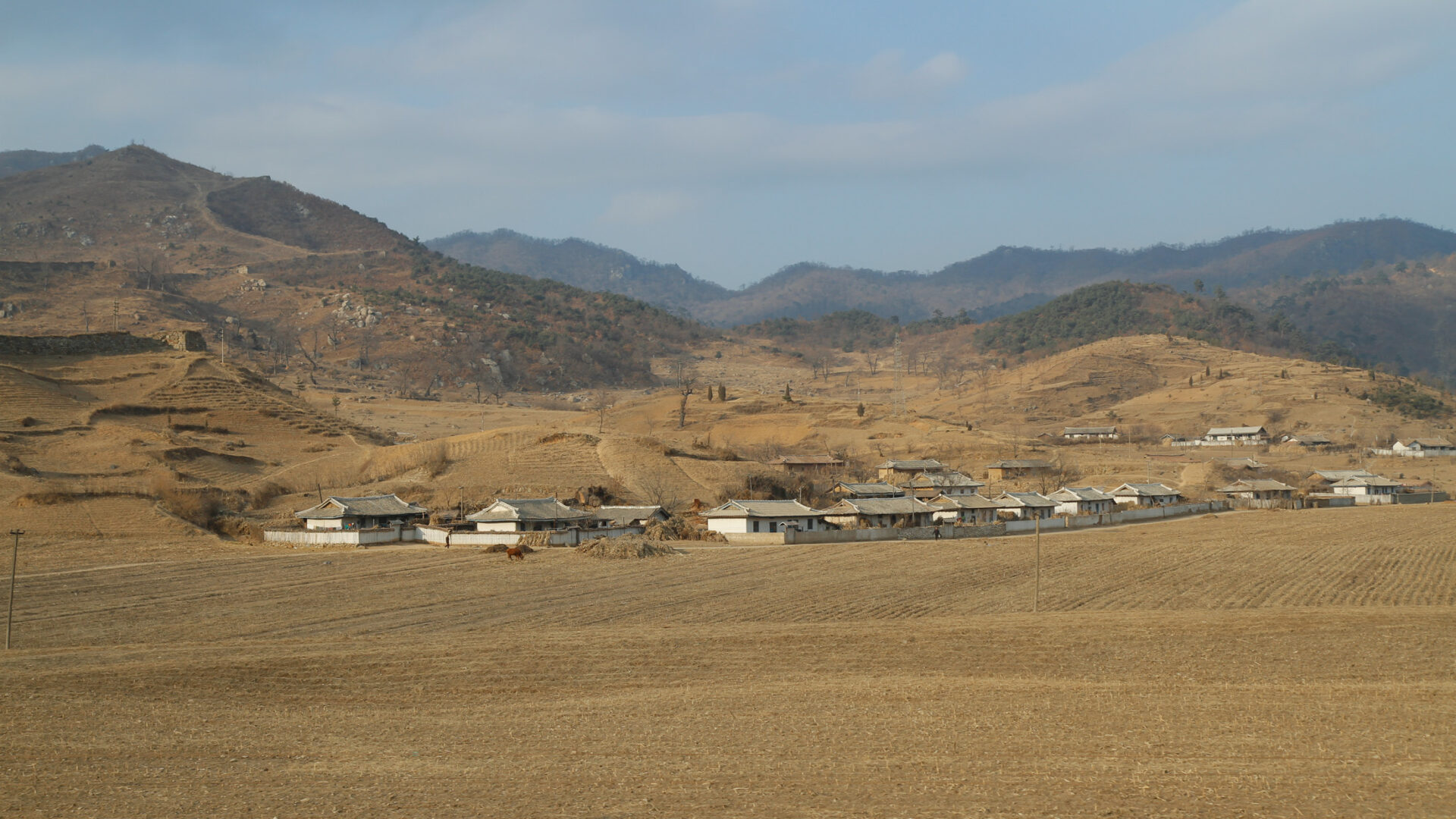 North Korean village surrounded by mountains, fields, and vegetation in a rugged landscape.
