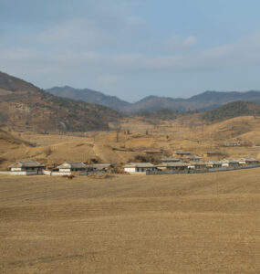 North Korean village surrounded by mountains, fields, and vegetation in a rugged landscape.