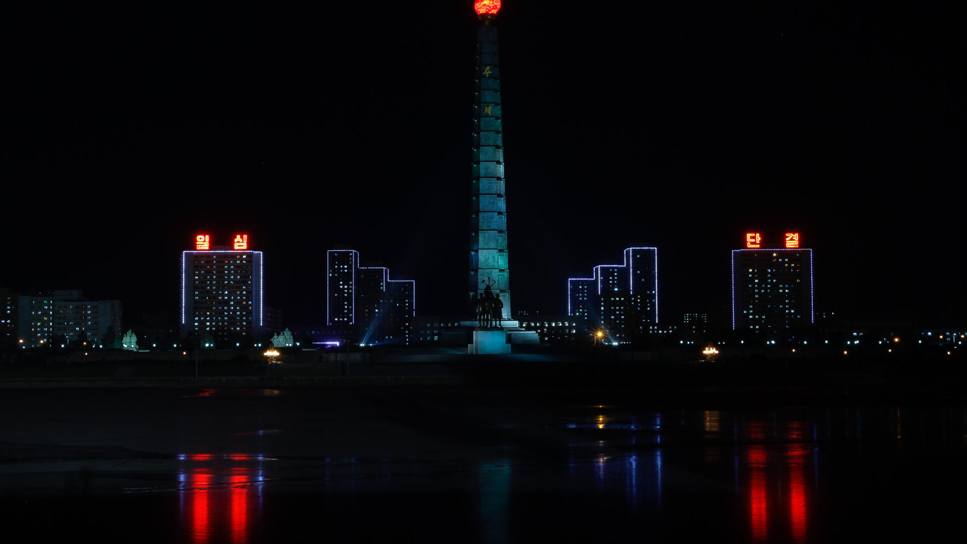 Pyongyang skyline at night with illuminated buildings and tower reflecting in still waters.