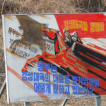 North Korea propaganda poster: soldier in action with red flag and patriotic text.