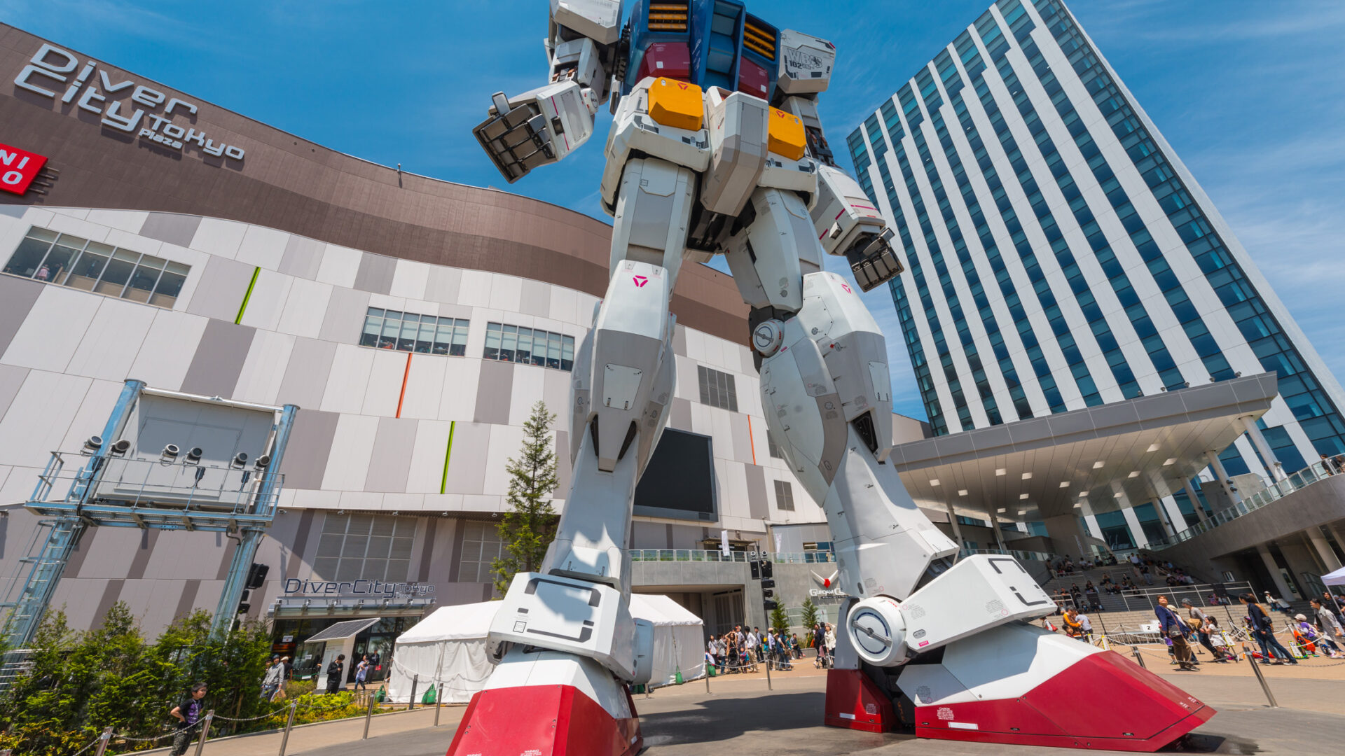 Impressive RX-78-2 Gundam statue in urban setting with bustling crowd and modern buildings.