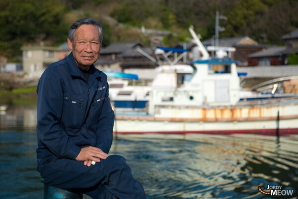 Tranquil scene of elderly man gazing at traditional boats in serene Manabe-shima, Japan.