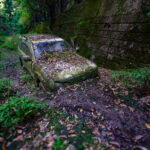 Abandoned Toyota Land Cruiser covered in moss, stuck in muddy forest beside stone wall