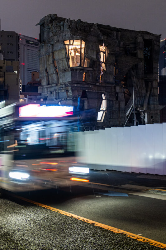 Ruined concrete building with lit windows as blurred bus passes at night city street.