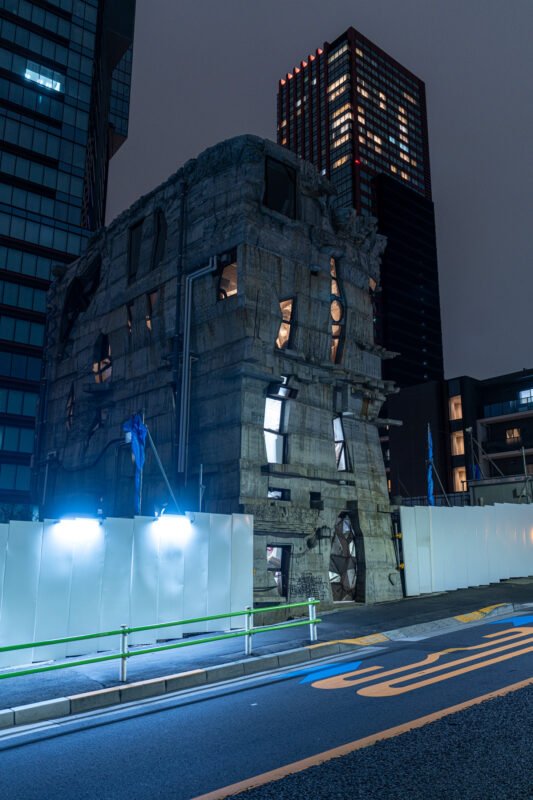 Partially demolished concrete building at night with construction lights and modern skyscrapers behind.