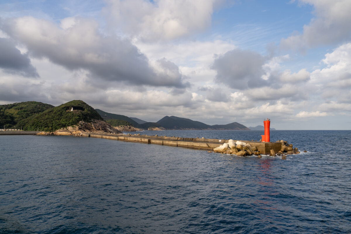 Red lighthouse on Tsushima breakwater overlooking calm blue sea and distant green hills.