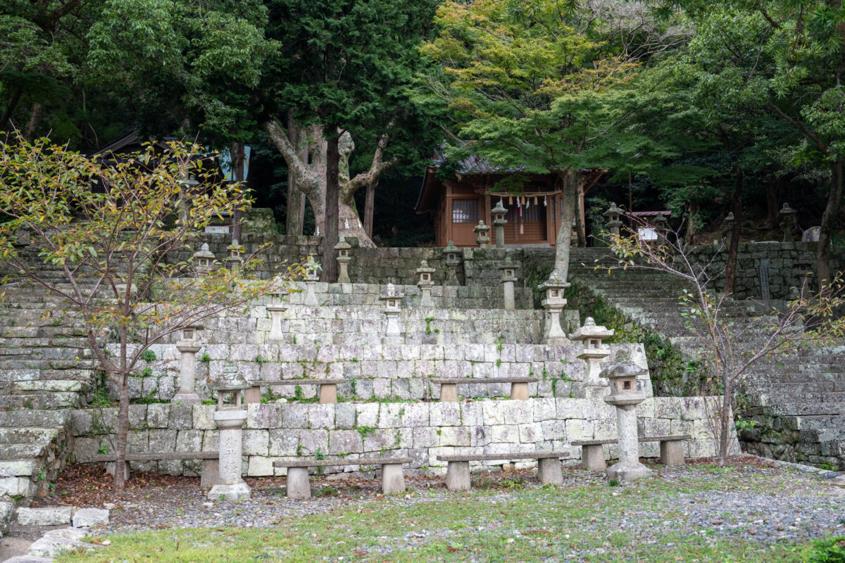 Terraced stone wall with lanterns leading to a forest shrine in Izuhara, Japan