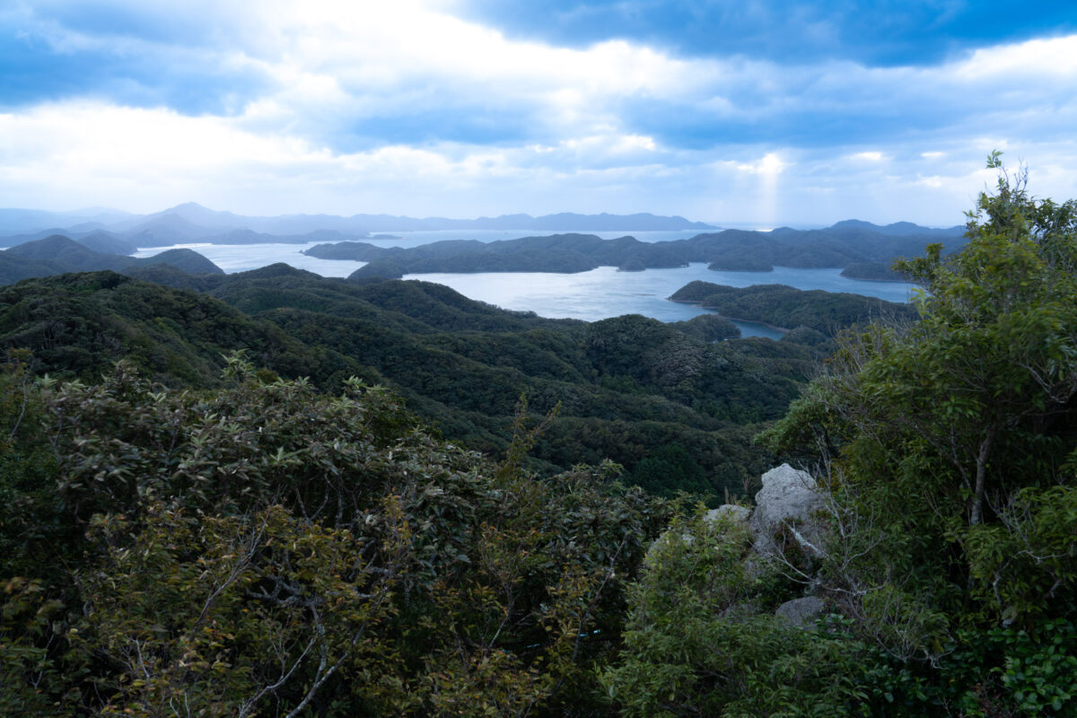 Panoramic Mount Eboshi summit view over coastal inlets and layered mountain ridges under cloudy skies.