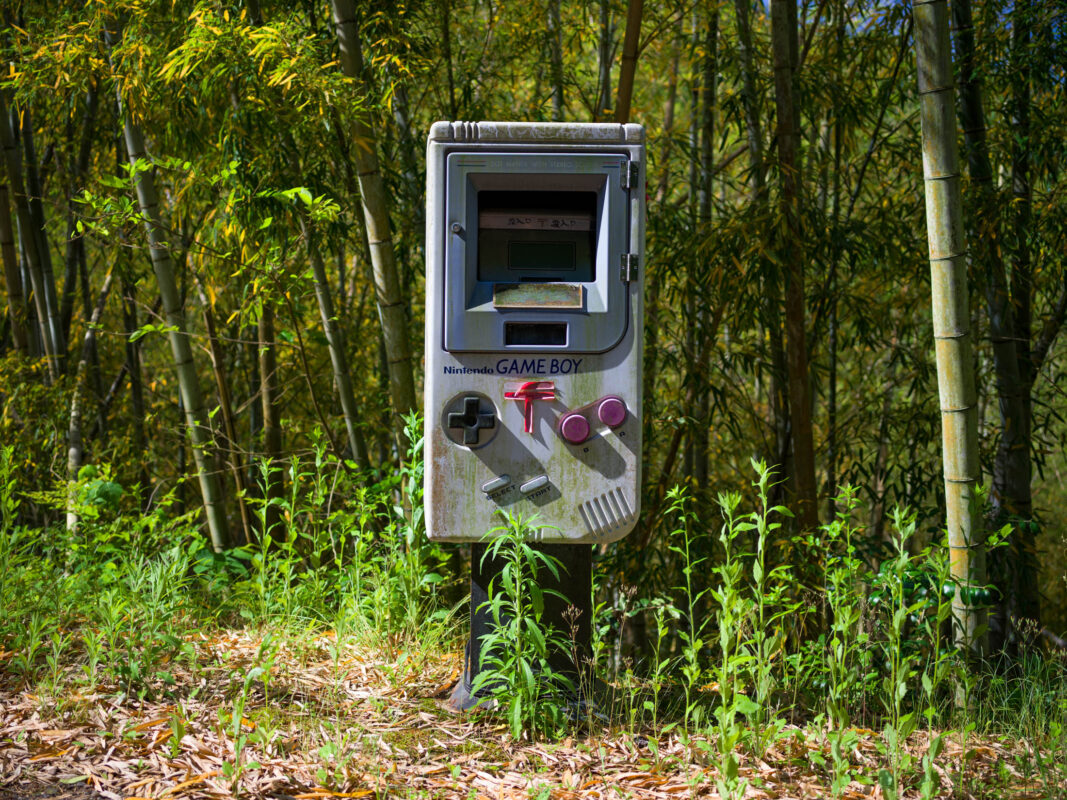 Weathered Game Boy-shaped mailbox on a post in a sunlit bamboo grove
