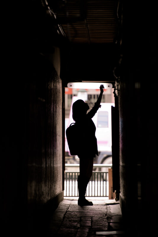 Silhouetted traveler with backpack raises arm in narrow Kyoto alley toward bright street