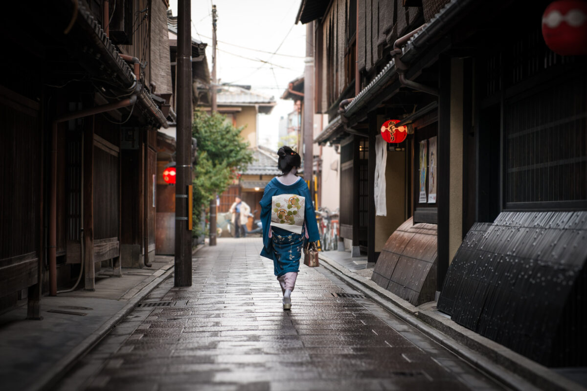 Person in blue kimono walking Kyoto stone alley with red lanterns after rain.