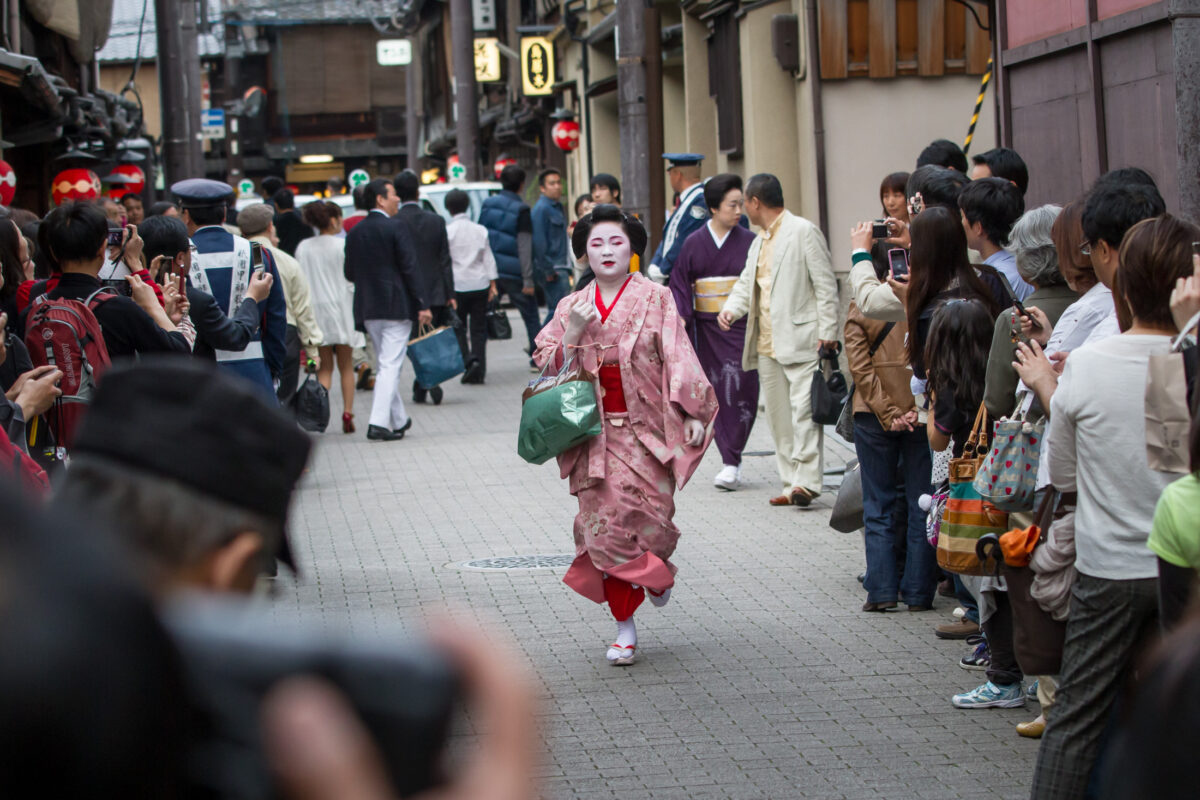 Geisha in pink kimono hurries down Kyoto street as tourists photograph her.