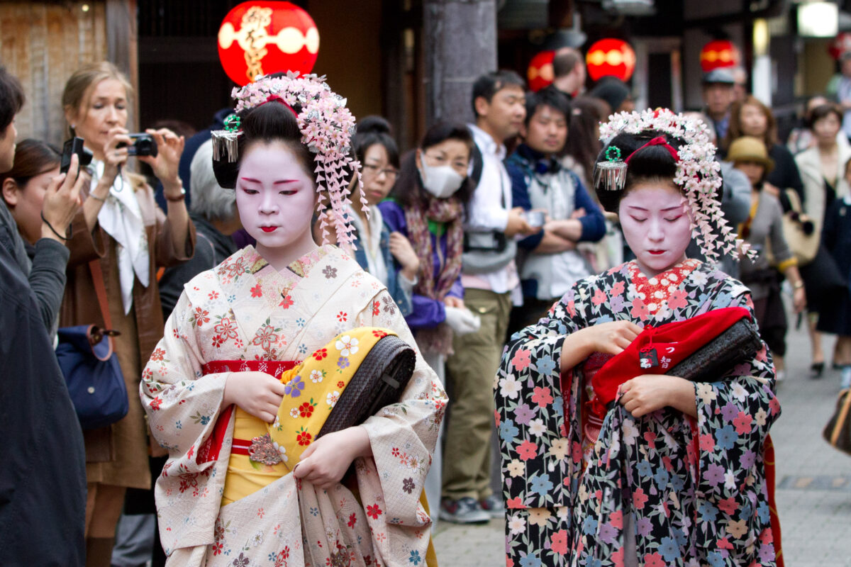 Two geisha in colorful kimono walk through Kyoto street crowd as tourists photograph them.