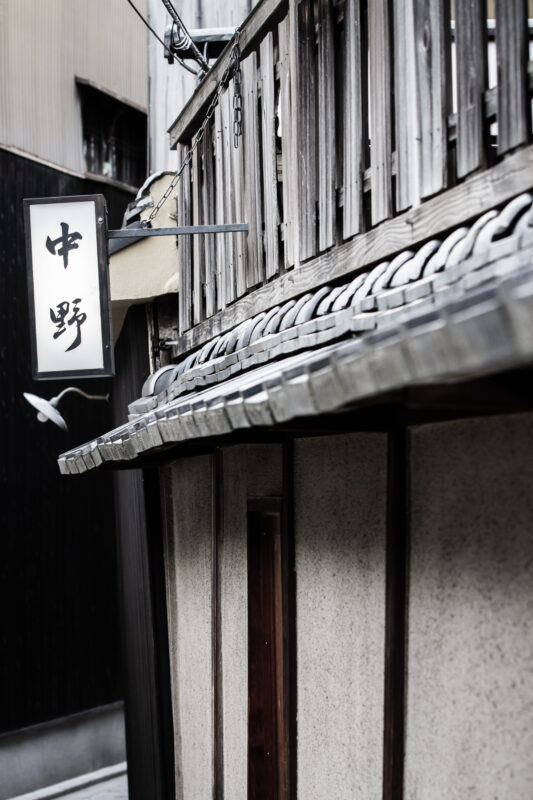 Traditional Japanese alley with weathered wooden facade, tiled eaves, and hanging kanji sign.